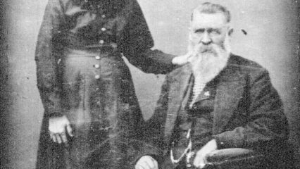 Black and white photo of Virgina Earp standing beside Nick Earp as he’s seated in a chair. They’re dressed in 19th century attire.