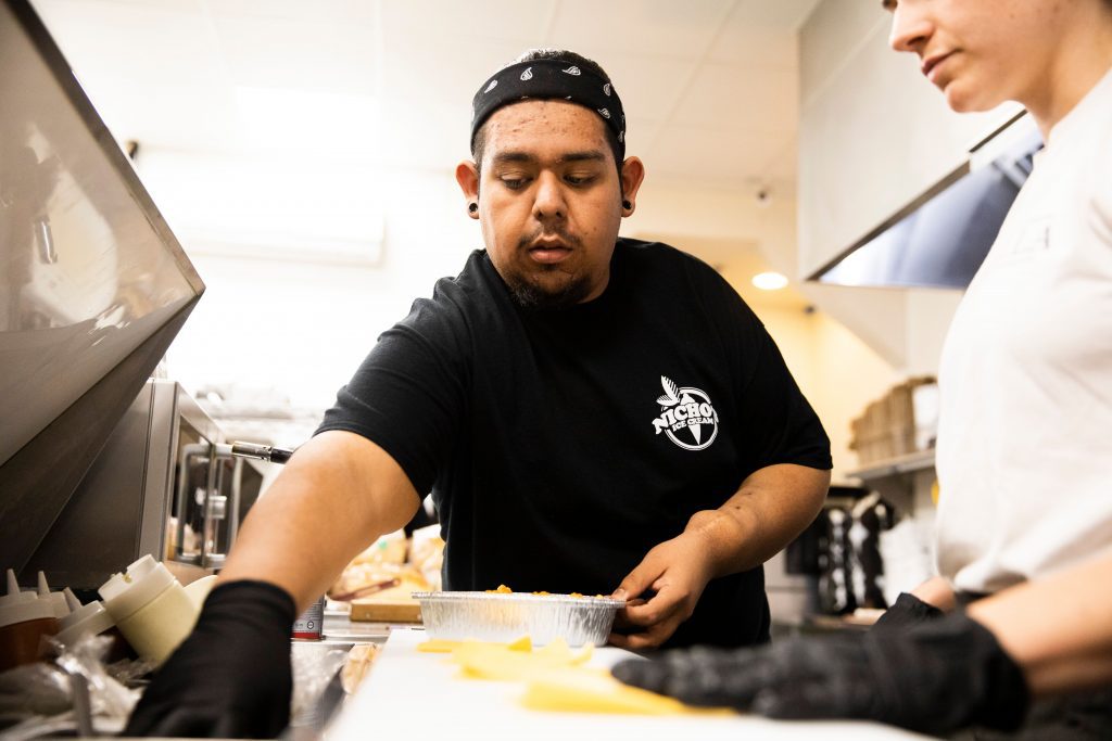 A male staff member at Nicho’s Ice Cream in Redlands prepares food in the kitchen, wearing a black Nicho’s Ice Cream shirt and bandana while focusing on the task.