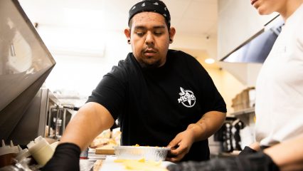 A male staff member at Nicho’s Ice Cream in Redlands prepares food in the kitchen, wearing a black Nicho’s Ice Cream shirt and bandana while focusing on the task.