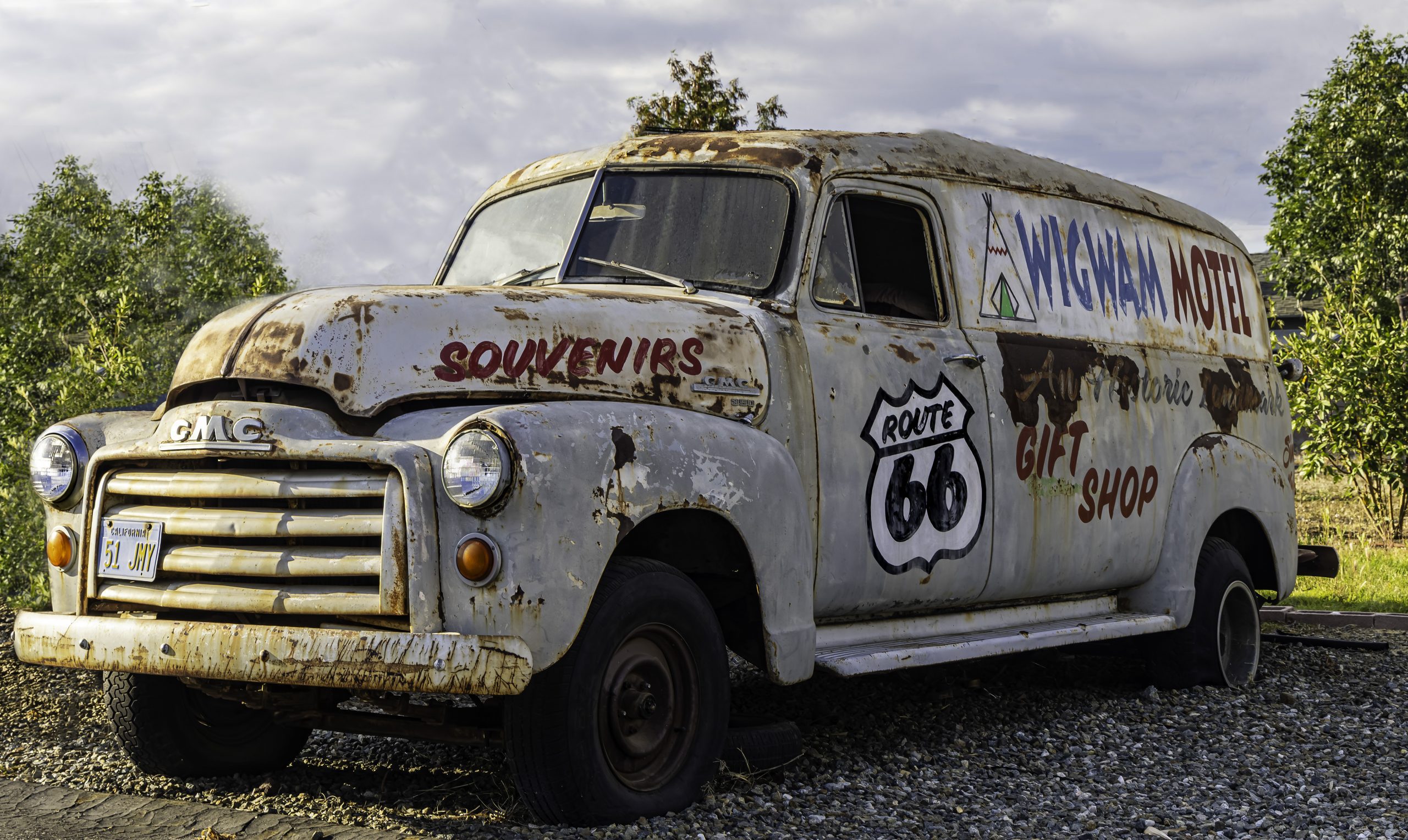 A weathered vintage GMC van is parked on a gravel surface surrounded by trees. The van displays faded signs reading "Wigwam Motel," "Route 66," "Souvenirs," and "Gift Shop."