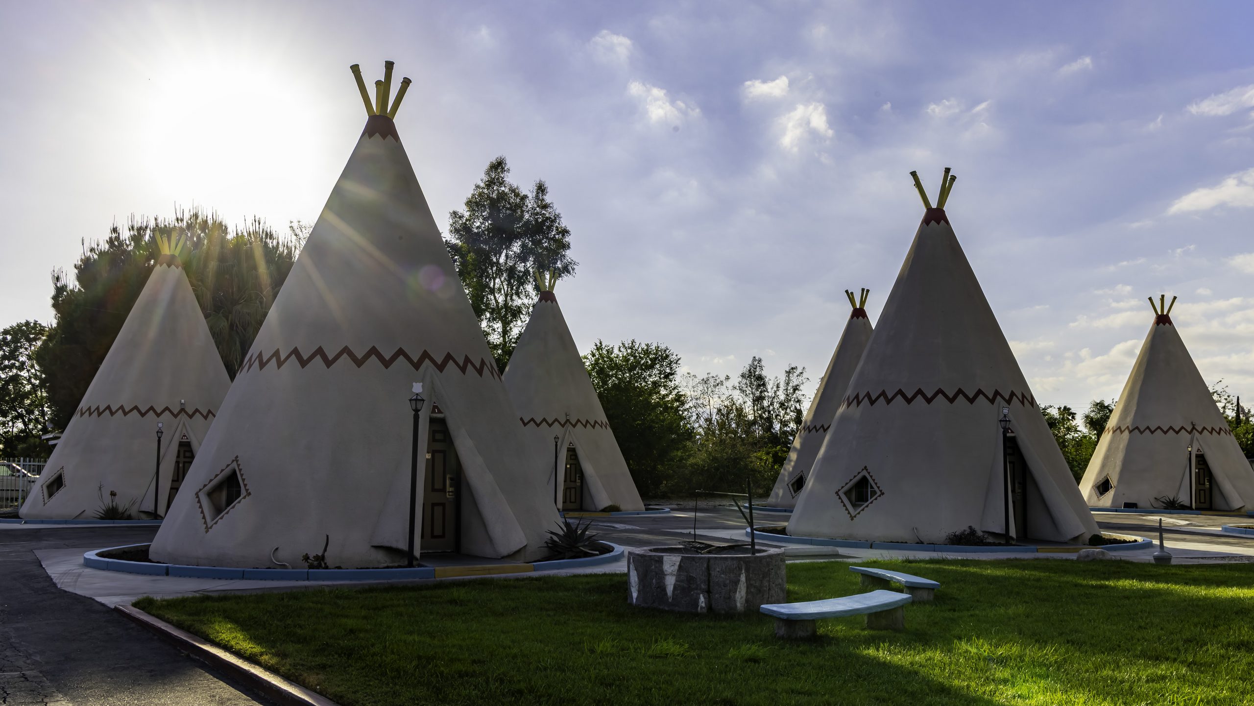 Four cone structures that resemble teepees under a partly cloudy sky. The sun is setting behind the trees, casting light over the scene.