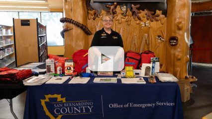 San Bernardino County Office of Emergency Services staff member stands behind a table displaying emer-gency preparedness supplies and information.