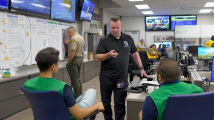 Staff coordinate disaster response in a command center, with a staff member briefing two team members in green vests while others work in the background.