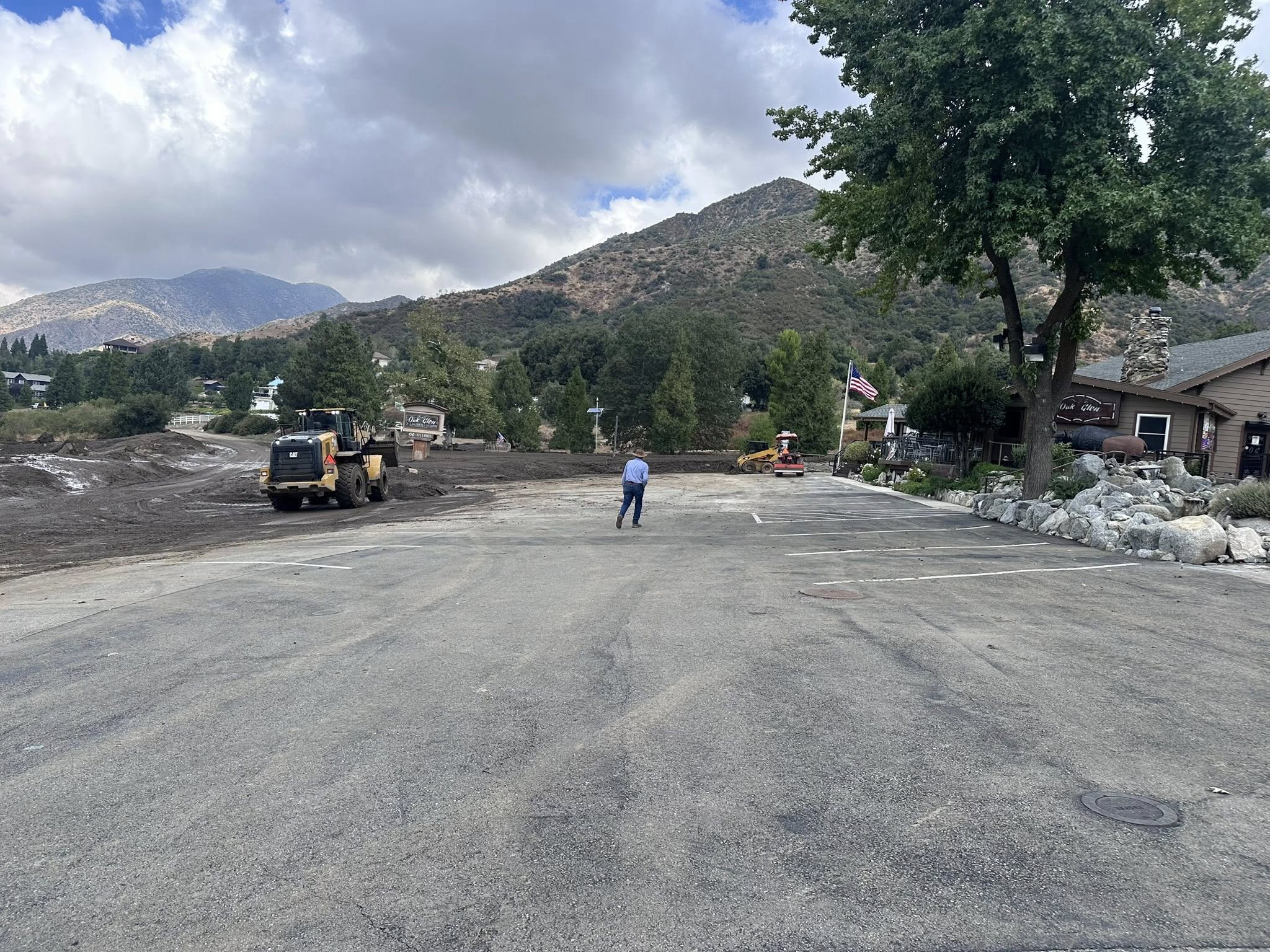 A person stands in a cleared parking lot as bulldozers move dirt nearby, with mountains and cloudy skies in the background.