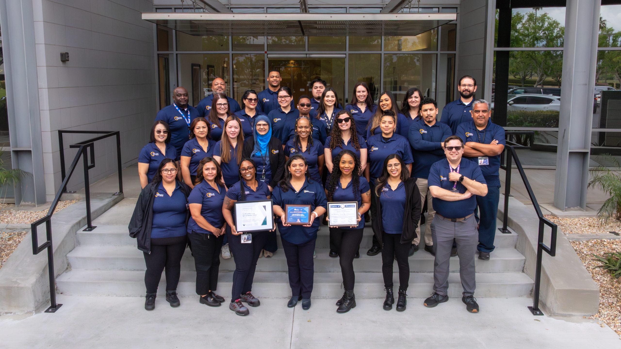 A group of people wearing matching polo shirts stand together on the steps in front of a building.