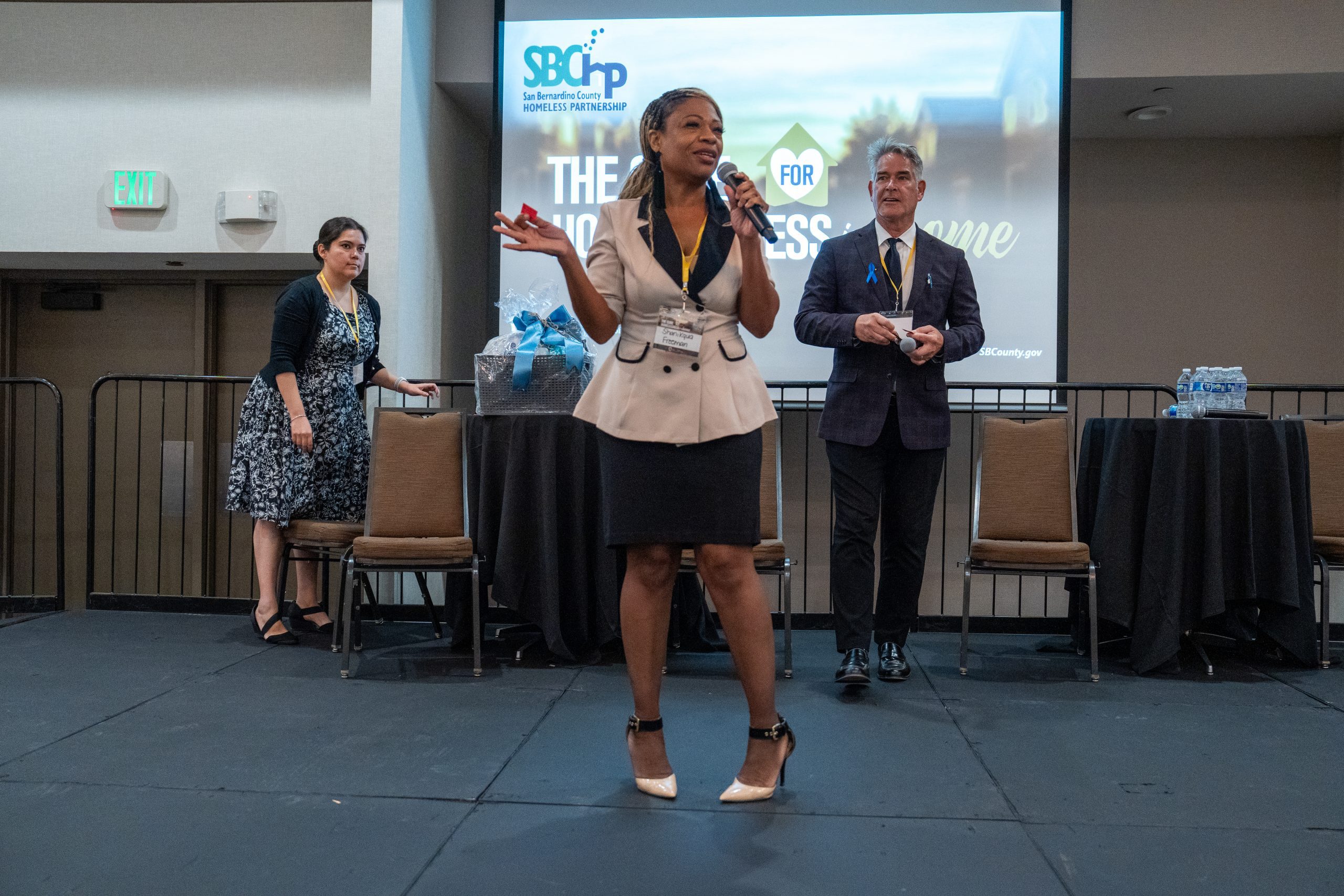 A woman in a pink suit is on stage speaking through a mic while a man in a suit is in the background.