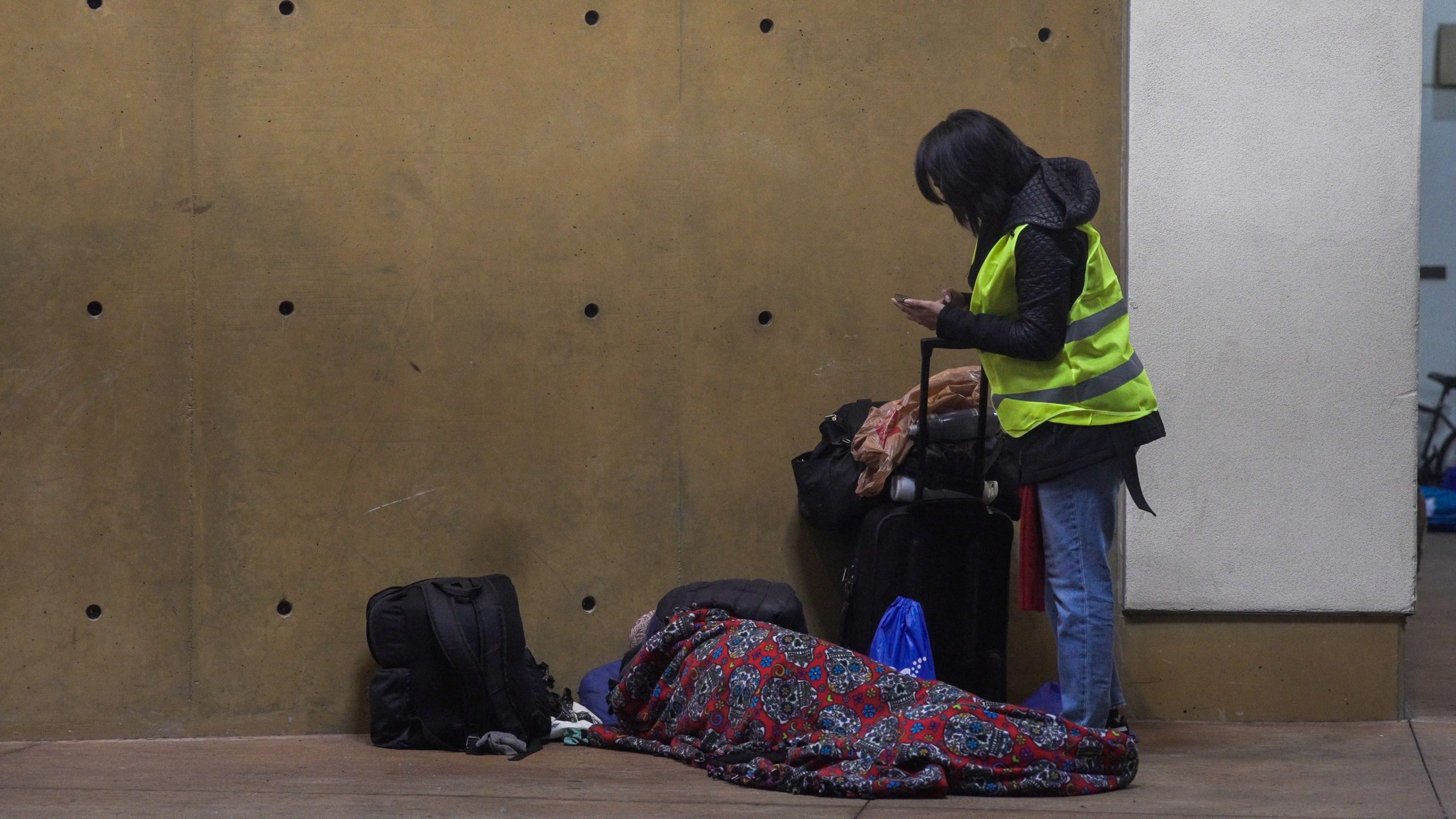 A person in a high visibility vest using a smartphone stands next to a person lying on the ground under a colorful blanket.