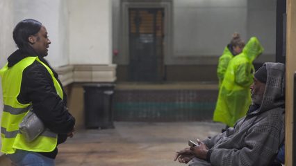 An individual in a safety vest speaks with a homeless person seated on a bench.