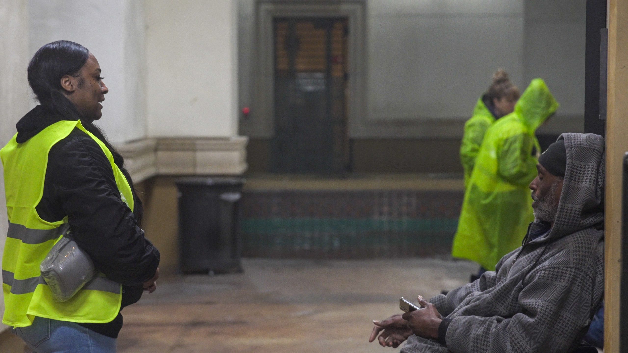 An individual in a safety vest speaks with a homeless person seated on a bench.