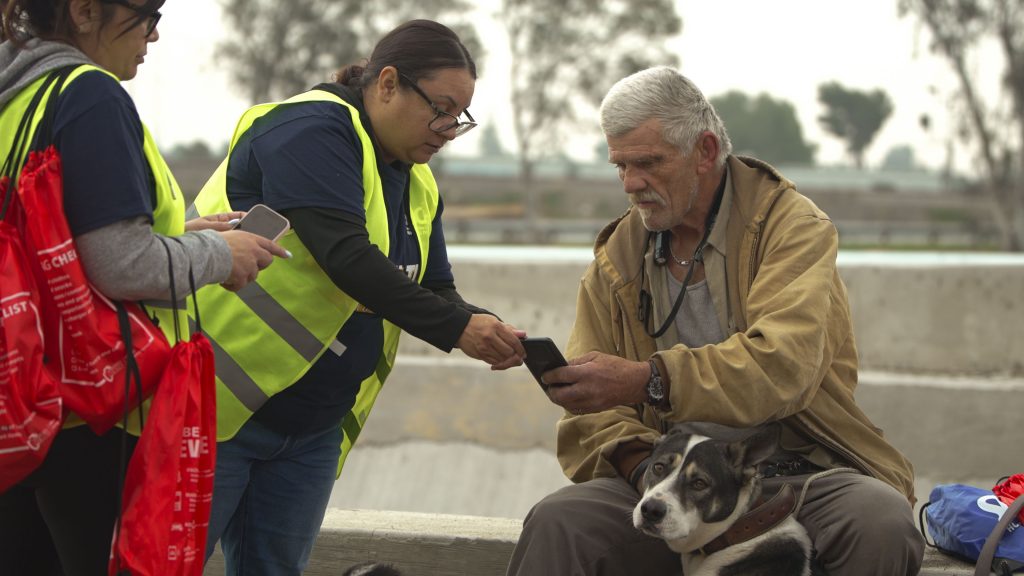Two people wearing reflective vests stand near an older individual with a dog. One person holds a smartphone towards the older individual.