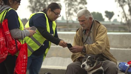 Two people wearing reflective vests stand near an older individual with a dog. One person holds a smartphone towards the older individual.