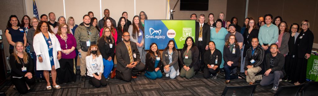 Group photo of diverse participants at a OneLegacy Donate Life event, smiling and standing around a banner with organization logos.