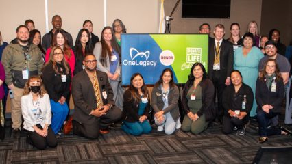 Group photo of diverse participants at a OneLegacy Donate Life event, smiling and standing around a banner with organization logos.
