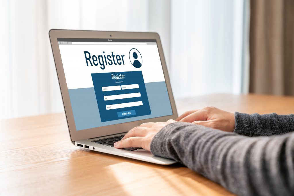A person types on a laptop. The screen displays a “Register” banner with various registration fields.