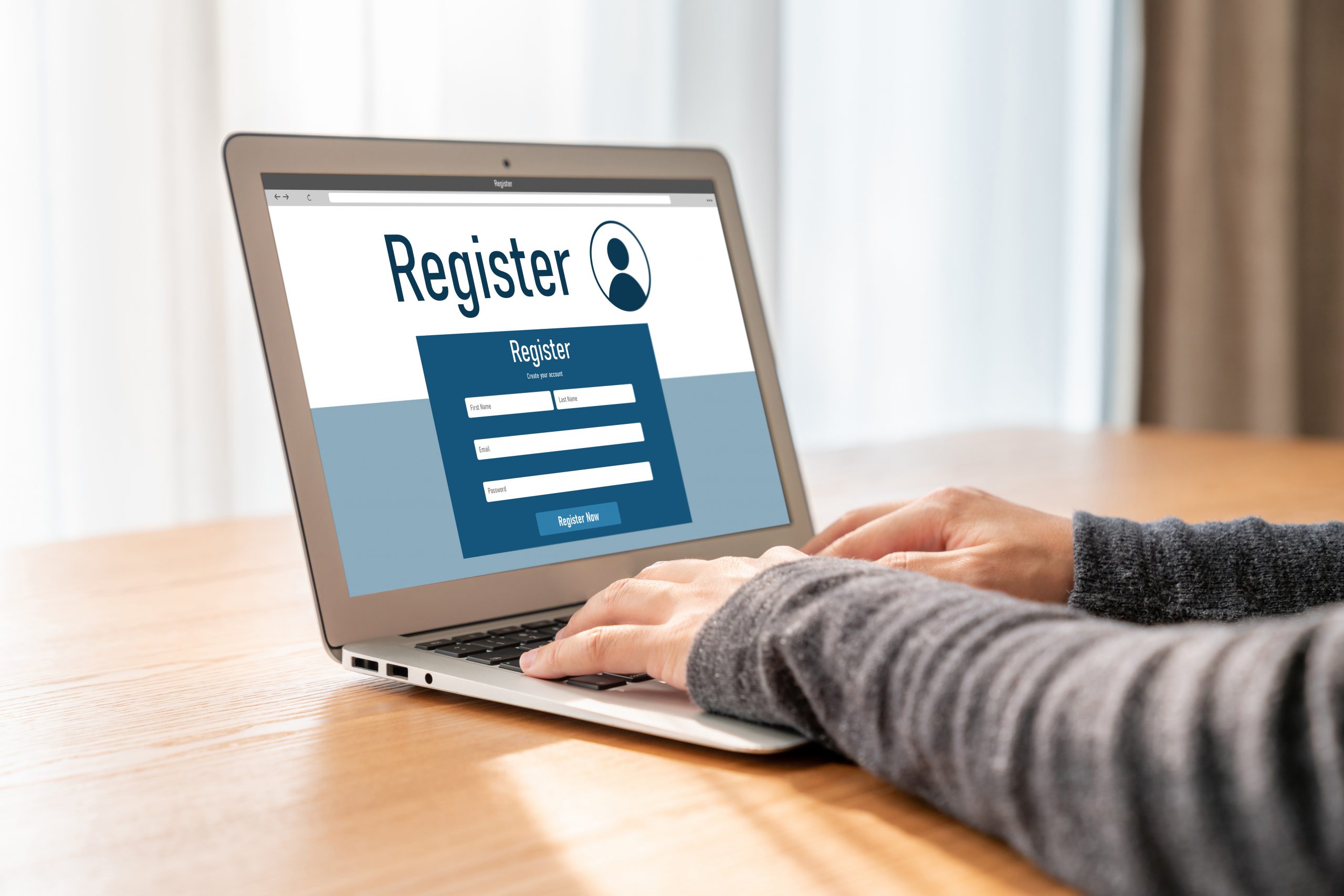 A person types on a laptop. The screen displays a “Register” banner with various registration fields.
