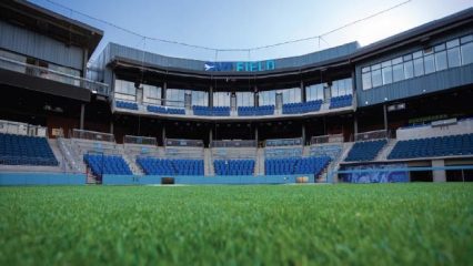 Low-angle view from the grass field inside an empty stadium with rows of blue seats, and the words “ONT FIELD” displayed above the concourse.