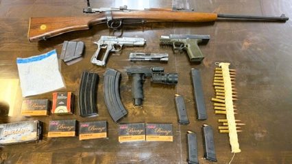 Top-down view of a wooden table displaying a bolt-action rifle, two handguns, magazines, loose ammo, and boxes of cartridges arranged neatly.
