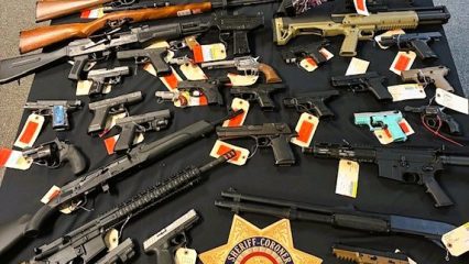 Table with rifles, shotguns, and handguns tagged as evidence; sheriff badge emblem; text: San Bernardino County Sheriff's Department, Sheriff-Coroner San Bernardino County.