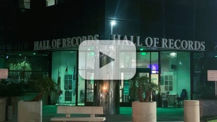 San Bernardino County Hall of Records building lit with green lights at night and a video play button; text on building reads “Hall of Records."