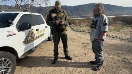 A San Bernardino County Sheriff’s Deputy speaks with an individual experiencing homelessness during 'Operation Shelter Me,' conducted by the Sheriff’s Homeless Outreach Proactive Enforcement (H.O.P.E.) Team. The interaction takes place outdoors in a rural area, with a Sheriff’s vehicle parked nearby and mountains visible in the background.