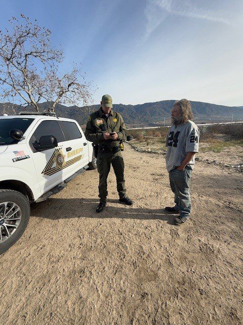 A San Bernardino County Sheriff’s Deputy speaks with an individual experiencing homelessness during 'Operation Shelter Me,' conducted by the Sheriff’s Homeless Outreach Proactive Enforcement (H.O.P.E.) Team. The interaction takes place outdoors in a rural area, with a Sheriff’s vehicle parked nearby and mountains visible in the background.