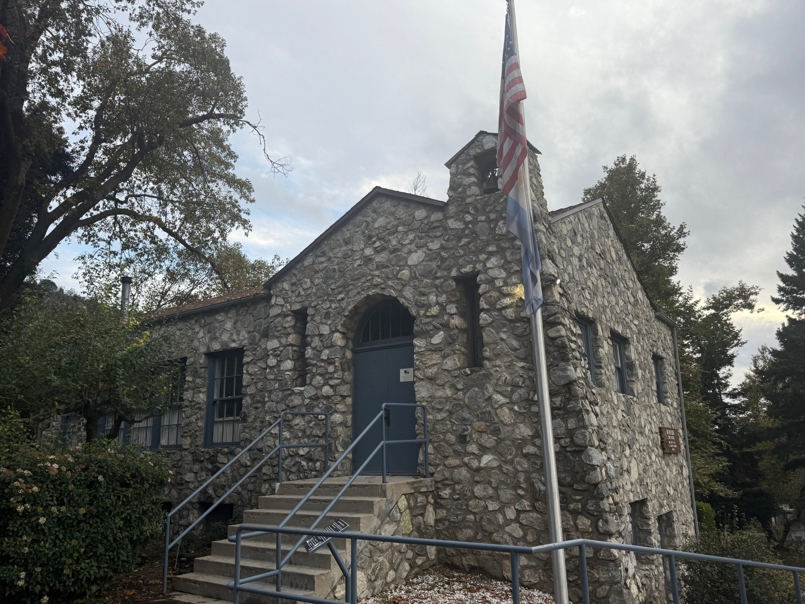 Exterior view of a stone building with a blue door. The American flag and another flag on a pole are displayed in front of the building.