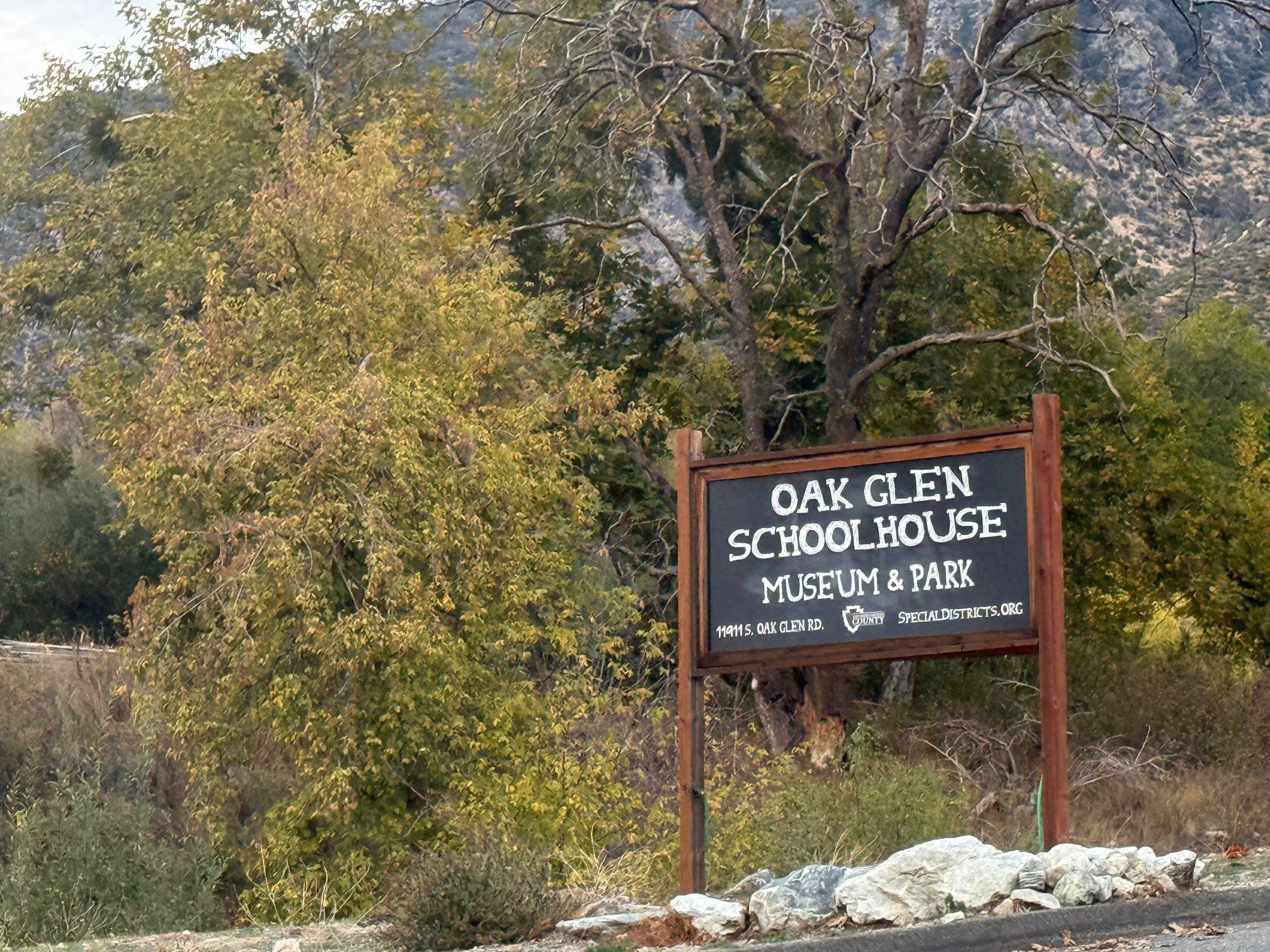 Sign for Oak Glen Schoolhouse Museum & Park in front of trees and a mountainous background.