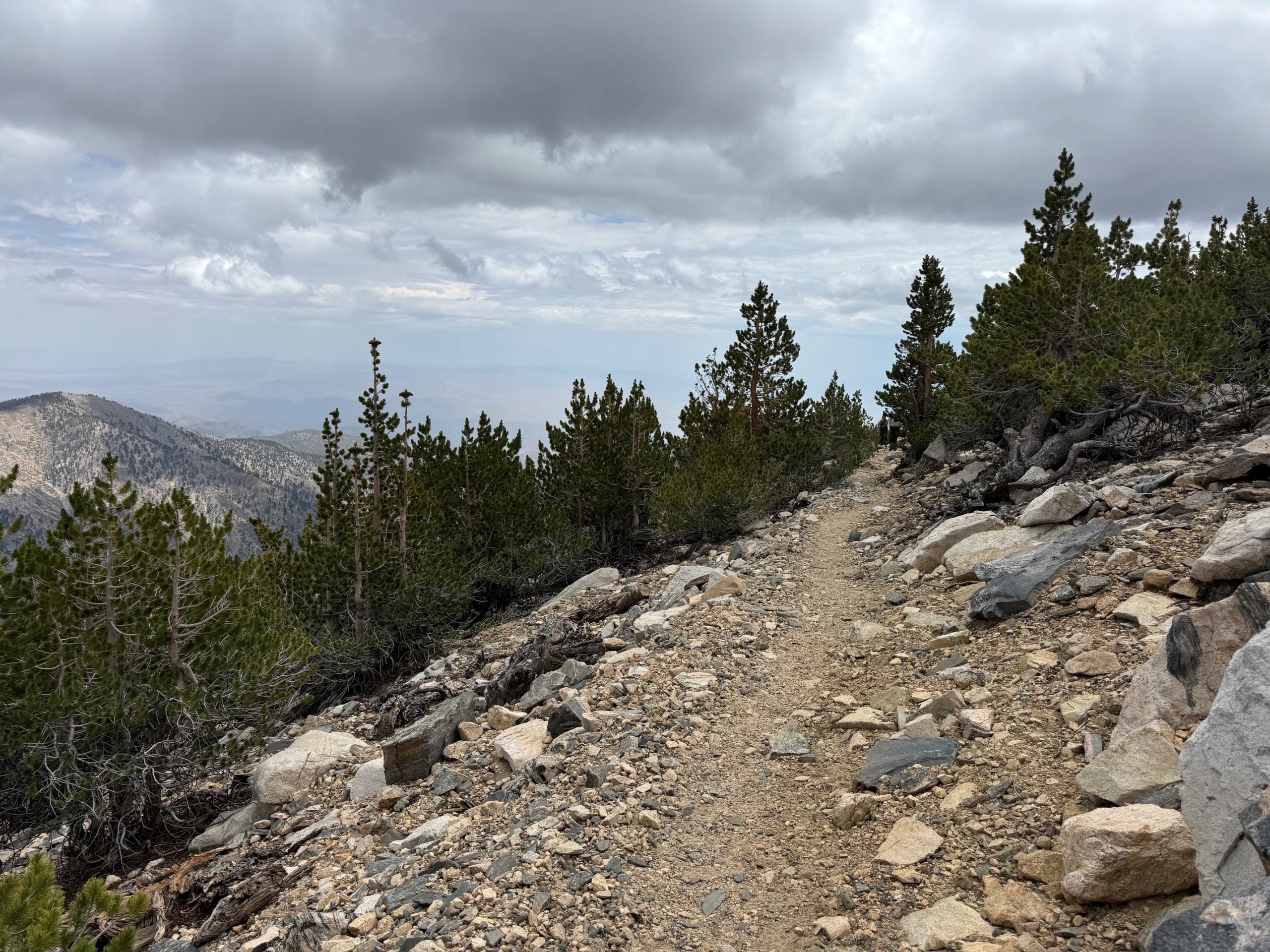 A trail that winds around a mountain under a partly cloudy sky surrounded by scattered trees and green shrubs.