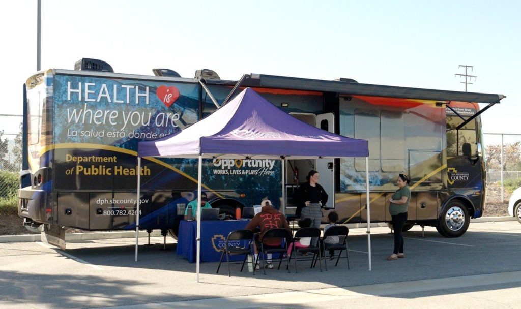 Mobile clinic in parking lot with purple pop-up tent in front and staff on a computer, speaking to visitors.
