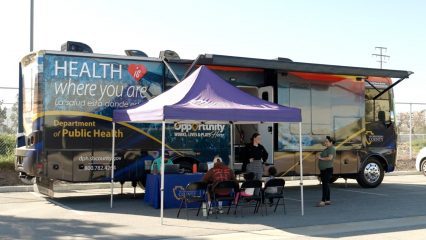 Mobile clinic in parking lot with purple pop-up tent in front and staff on a computer, speaking to visitors.