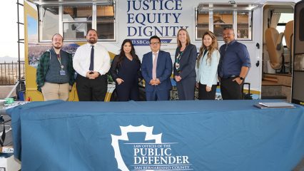 Seven mobile public defender team members standing behind a table. The table has the San Bernardino County Law Office of the Public Defender displayed. Behind the team is their mobile defense unit that states Law Offices of the Public Defender, Justice, Equity, and Dignity.