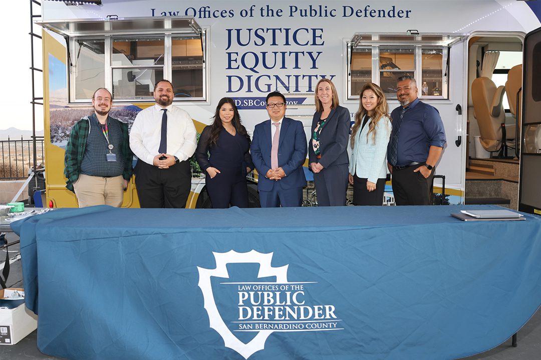 Seven mobile public defender team members standing behind a table. The table has the San Bernardino County Law Office of the Public Defender displayed. Behind the team is their mobile defense unit that states Law Offices of the Public Defender, Justice, Equity, and Dignity.