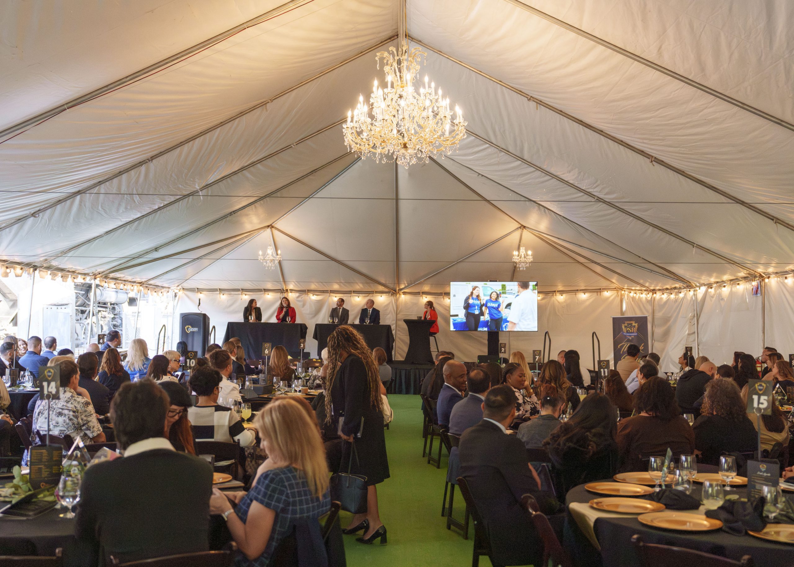 A large event inside a white tent features a seated audience facing a stage with five presenters standing at a long table. A screen displays a presentation. Tables are adorned with black tablecloths and place settings, and there is an elegant chandelier hanging from the tent ceiling.