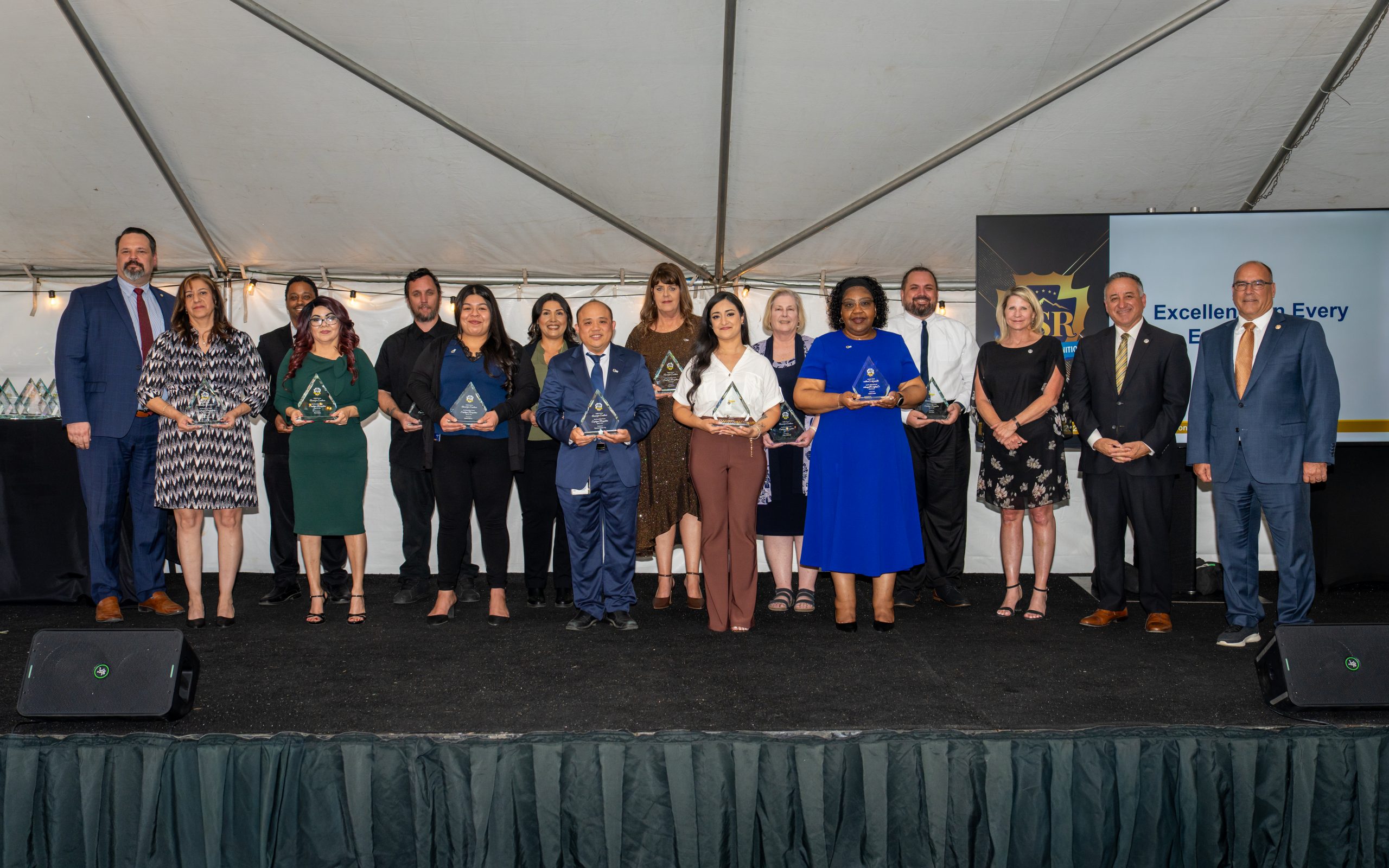 A group of people stands on a stage under a tent, holding various awards. All are dressed in formal or business attire, and there are smiles and expressions of accomplishment.