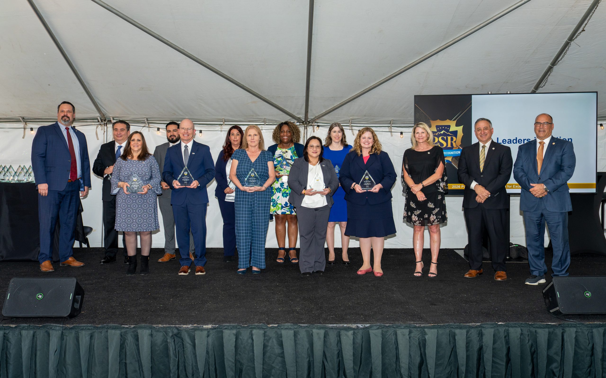 A group of individuals stands on a stage under a tent, each holding an award. A screen in the background displays "PSR" and "Leadership."