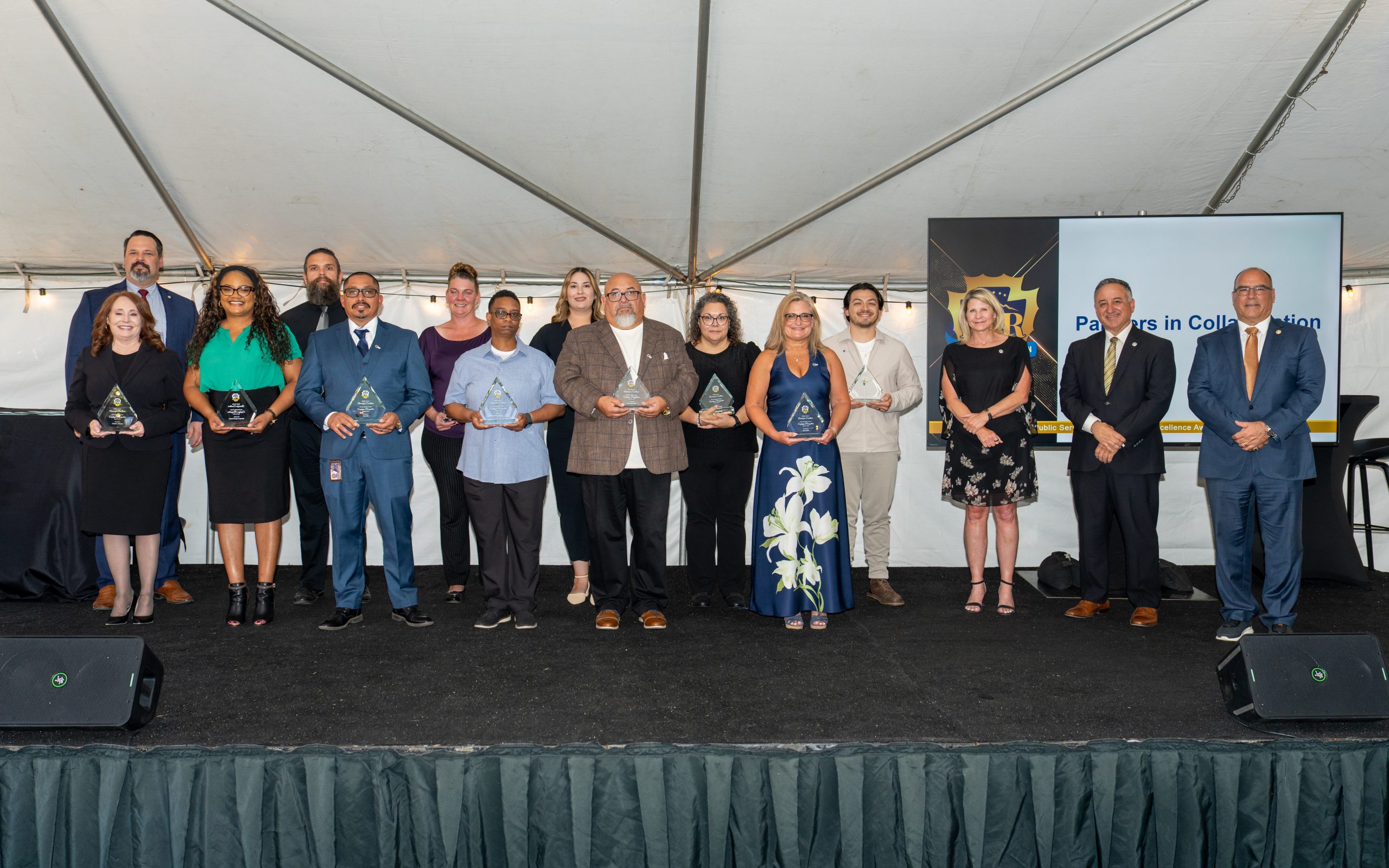A group of individuals stands on a stage under a tent, each holding an award. A screen in the background displays "PSR" and "Partners in Collaboration."