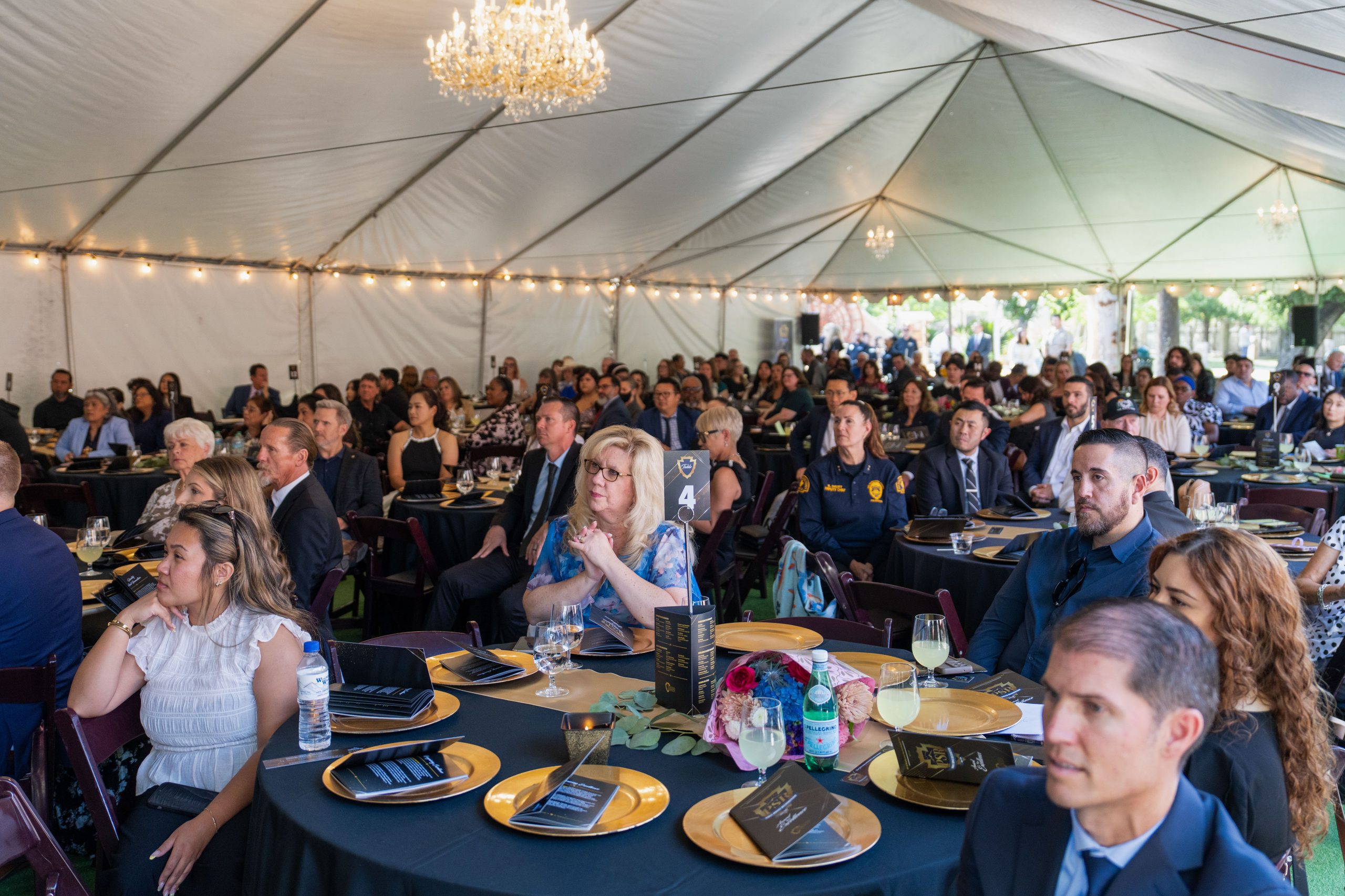 A large gathering takes place under a white event tent with chandeliers. Attendees are seated at round tables adorned with black tablecloths and gold plates. The audience appears attentive and engaged.
