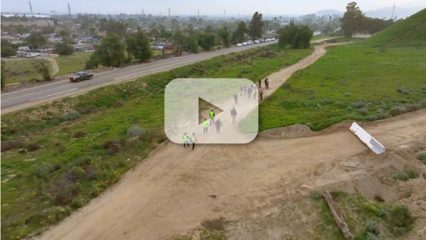 A group of people are walking on a dirt path near a road. There's a large play button overlaid on the image suggesting a video.