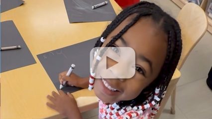 Smiling child with beaded braids looks up at the camera while drawing on black paper with a white crayon at a classroom table.