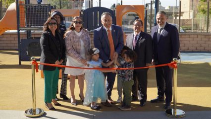 Six men and women in business attire and three children stand on a playground posing with one of the men holding large fake scissors to cut a red ribbon tied to two gold stanchions. Shadows of the individuals appear behind them.
