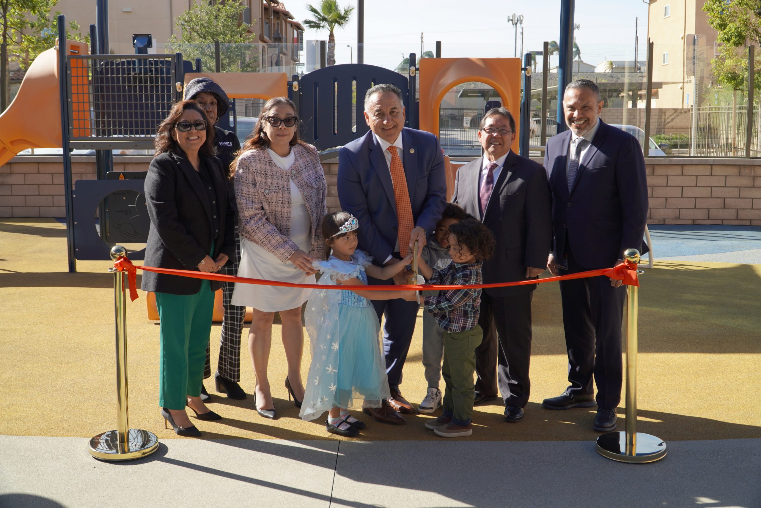 Six men and women in business attire and three children stand on a playground posing with one of the men holding large fake scissors to cut a red ribbon tied to two gold stanchions. Shadows of the individuals appear behind them.