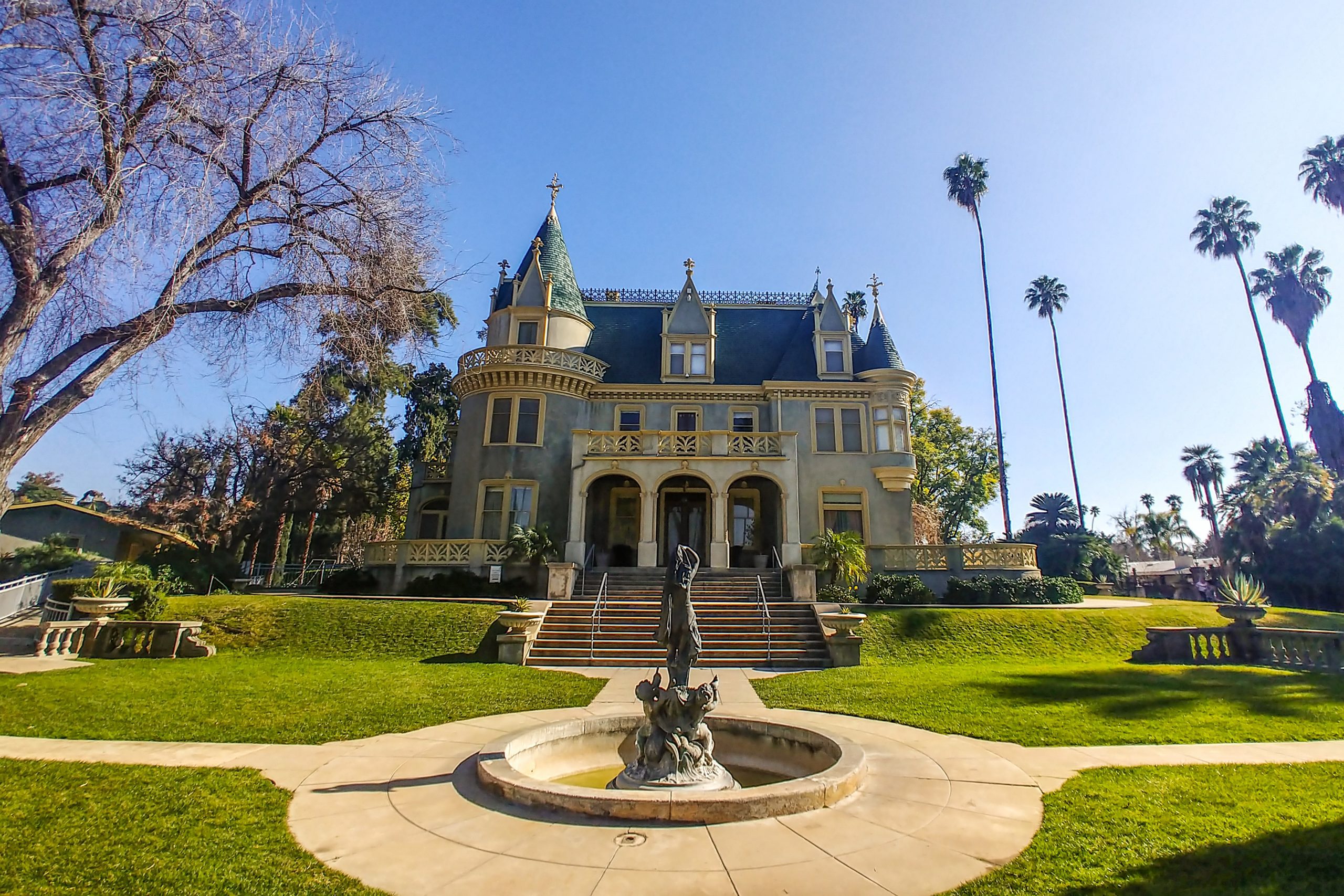 View of the front exterior of the Kimberly Crest House and Gardens featuring a large Victorian mansion surrounded by gardens and a water fountain in the foreground.