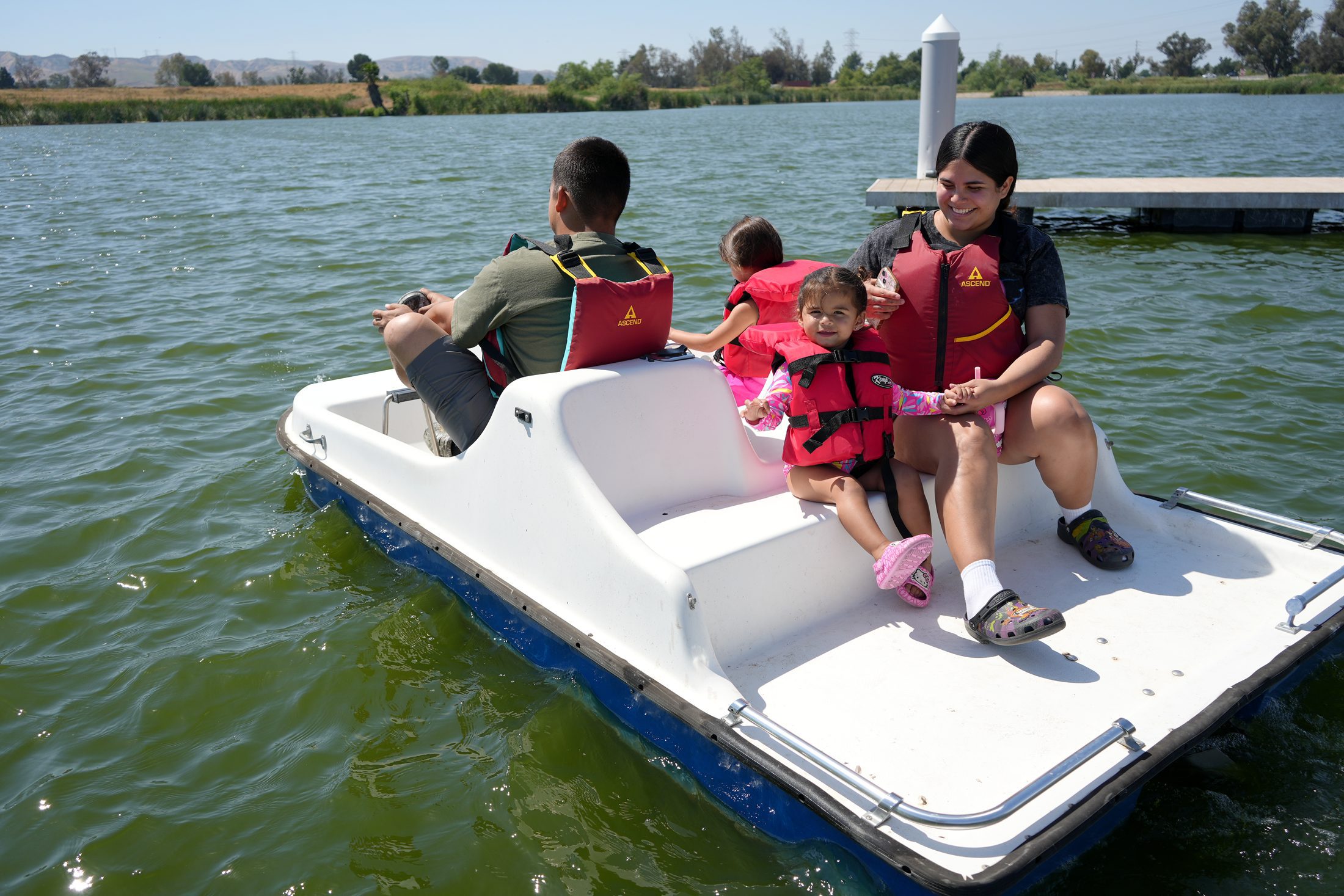A man and little girl sit in front of a pedal boat on a lake with a woman and a little girl sitting in the back.