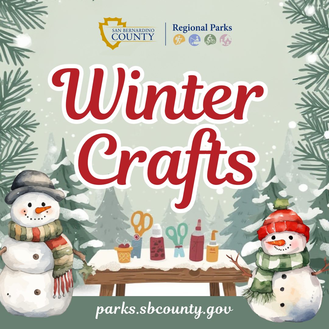 Two snowmen flank a picnic table with arts & crafts items covered in snow with trees and snow falling in the background.