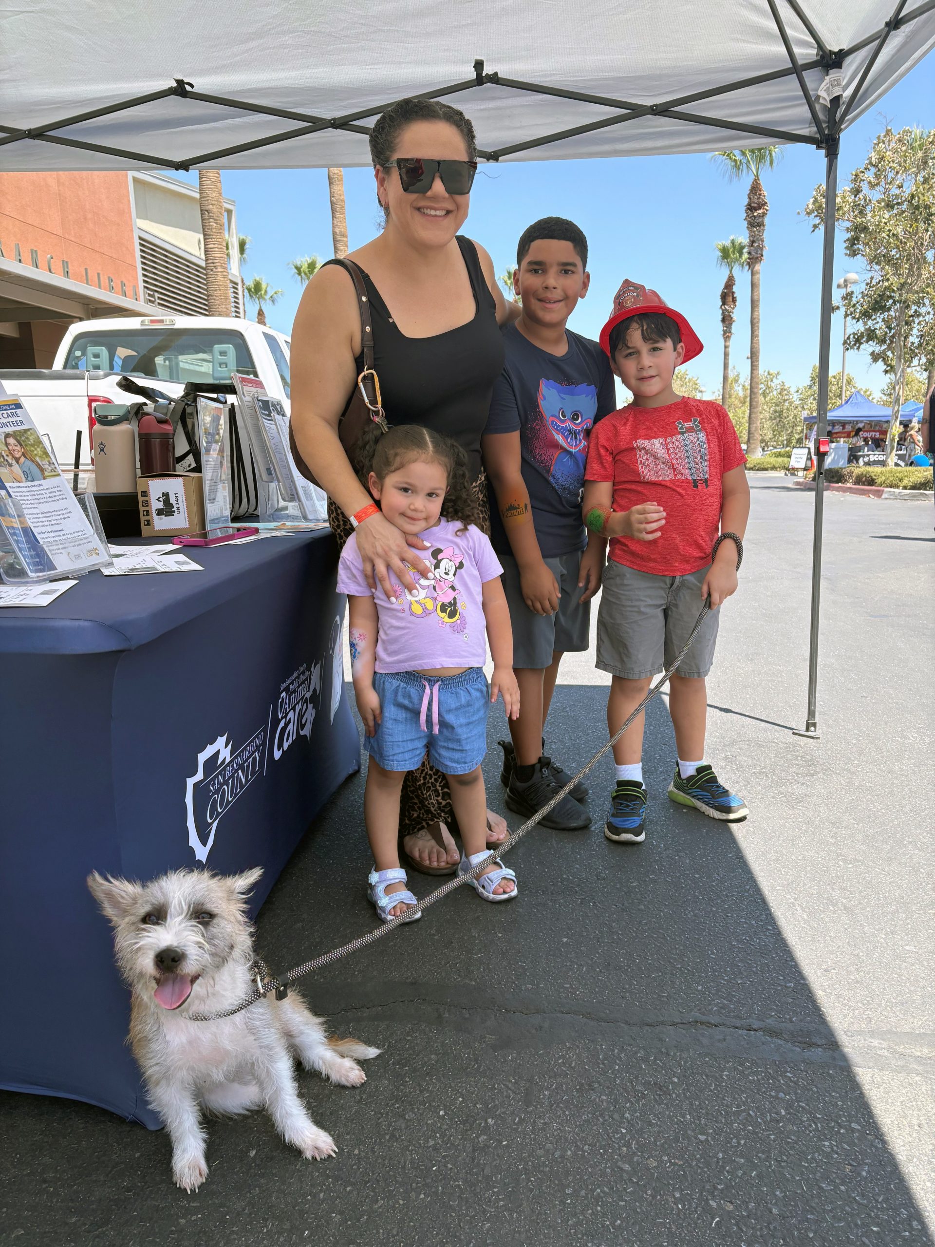 Smiling family of four poses beside a community booth with a small dog in front, under a sunny outdoor canopy; cheerful festival atmosphere.