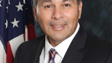 A man in a dark suit and striped tie smiles in a formal portrait, standing beside a U.S. flag against a blue studio backdrop.