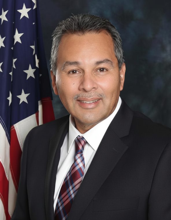 A man in a dark suit and striped tie smiles in a formal portrait, standing beside a U.S. flag against a blue studio backdrop.