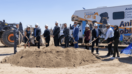 Group in business attire and hard hats pose with shovels at a groundbreaking site, with an Animal Care vehicle and bulldozer in the background.