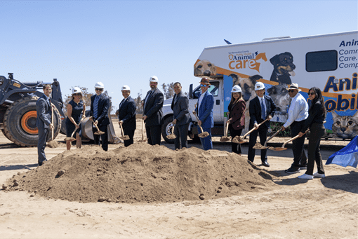 Group in business attire and hard hats pose with shovels at a groundbreaking site, with an Animal Care vehicle and bulldozer in the background.