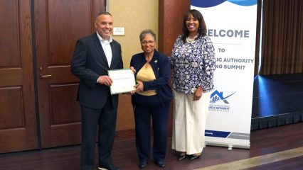 A play button icon for a video placed over a photo of a man in a dark suit and a woman in white pants and a purple and white top standing in front of an IHSS Public Authority Caregiver Training Summit banner, presenting a certificate to a woman in a navy suit.